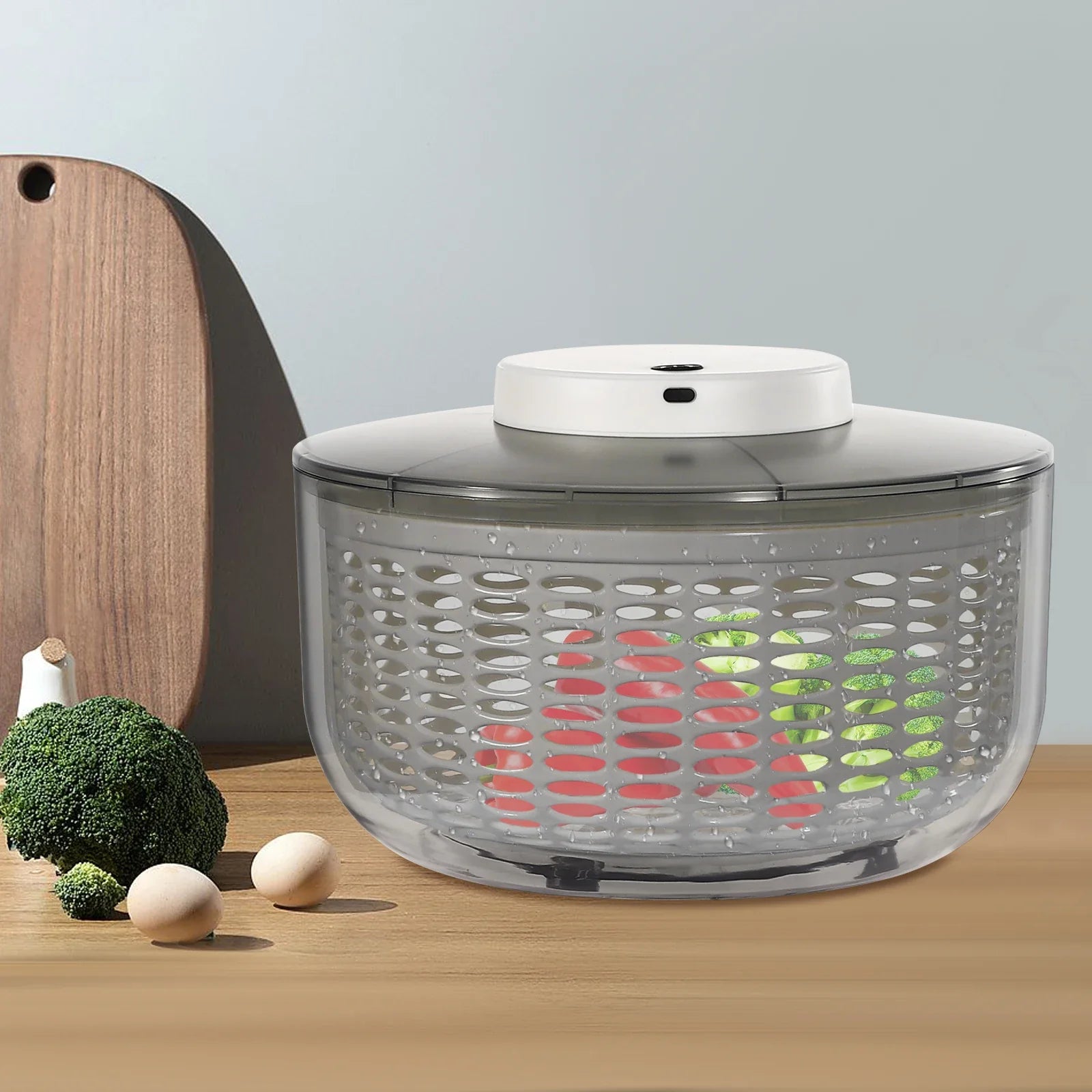 A salad spinner with a clear plastic basket and a white top button, shown on a wooden table with broccoli, eggs, and a cutting board in a kitchen setting.