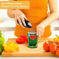 A person in an orange top uses a black electric can opener to open a green can with a red tomato graphic label, set on a wooden cutting board with fresh vegetables in a bright kitchen.
