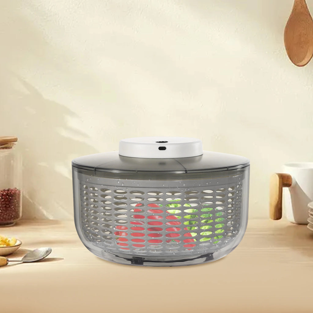 A countertop salad spinner with a clear plastic basket, dark gray lid, and white top button, shown on a beige kitchen counter with vegetables inside.