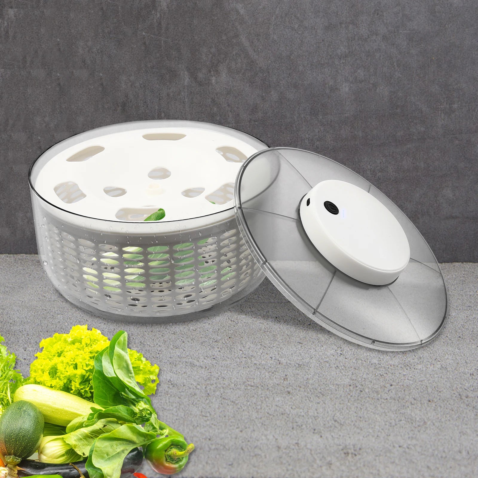 A clear plastic salad spinner basket with a white inner spinning plate and a matching clear lid with a white knob, shown on a gray surface with vegetables in the foreground.