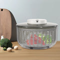 A salad spinner with a clear plastic basket and a white top button, shown on a wooden table with broccoli, eggs, and a cutting board in a kitchen setting.