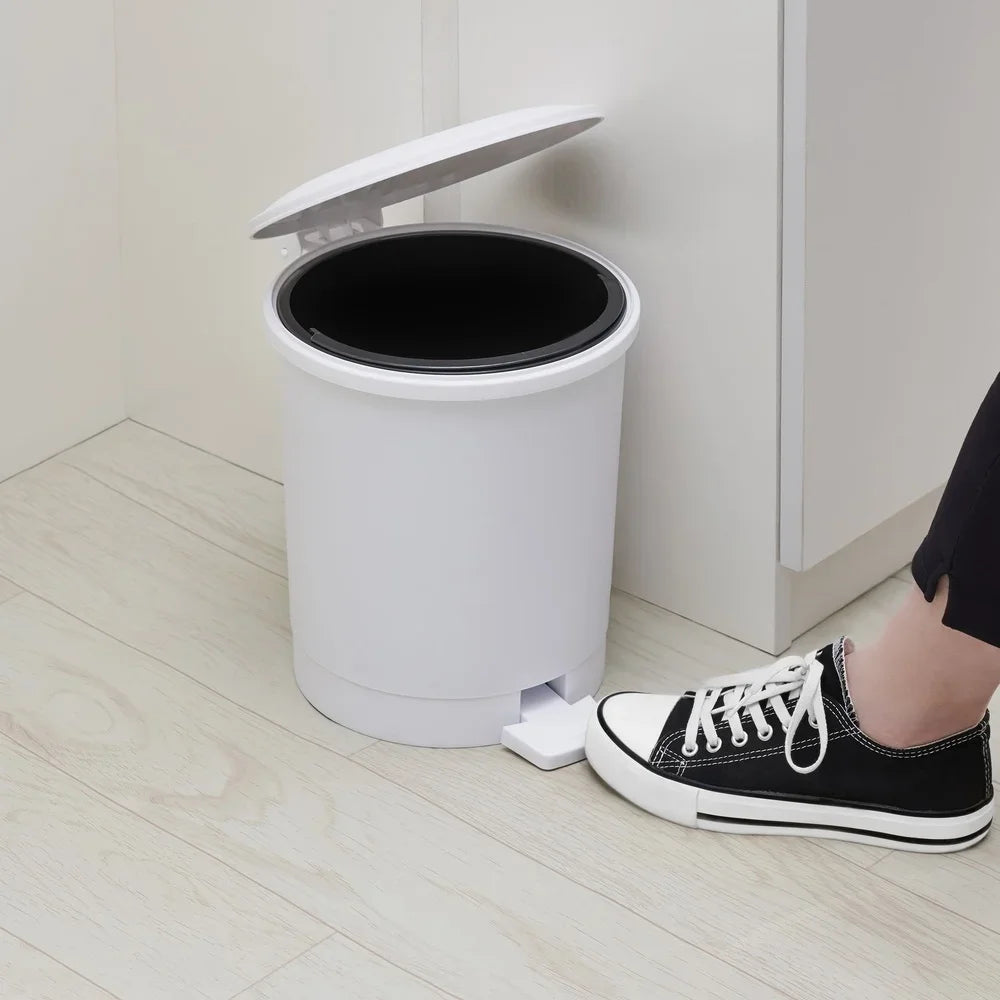 White trash bin with foot pedal on a wooden floor next to a person wearing black sneakers.