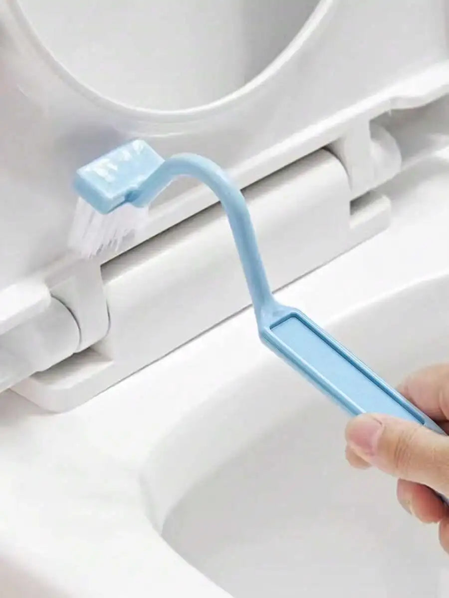 A hand holding a light blue toilet brush with white bristles, cleaning the hinge area of a white toilet seat and bowl.