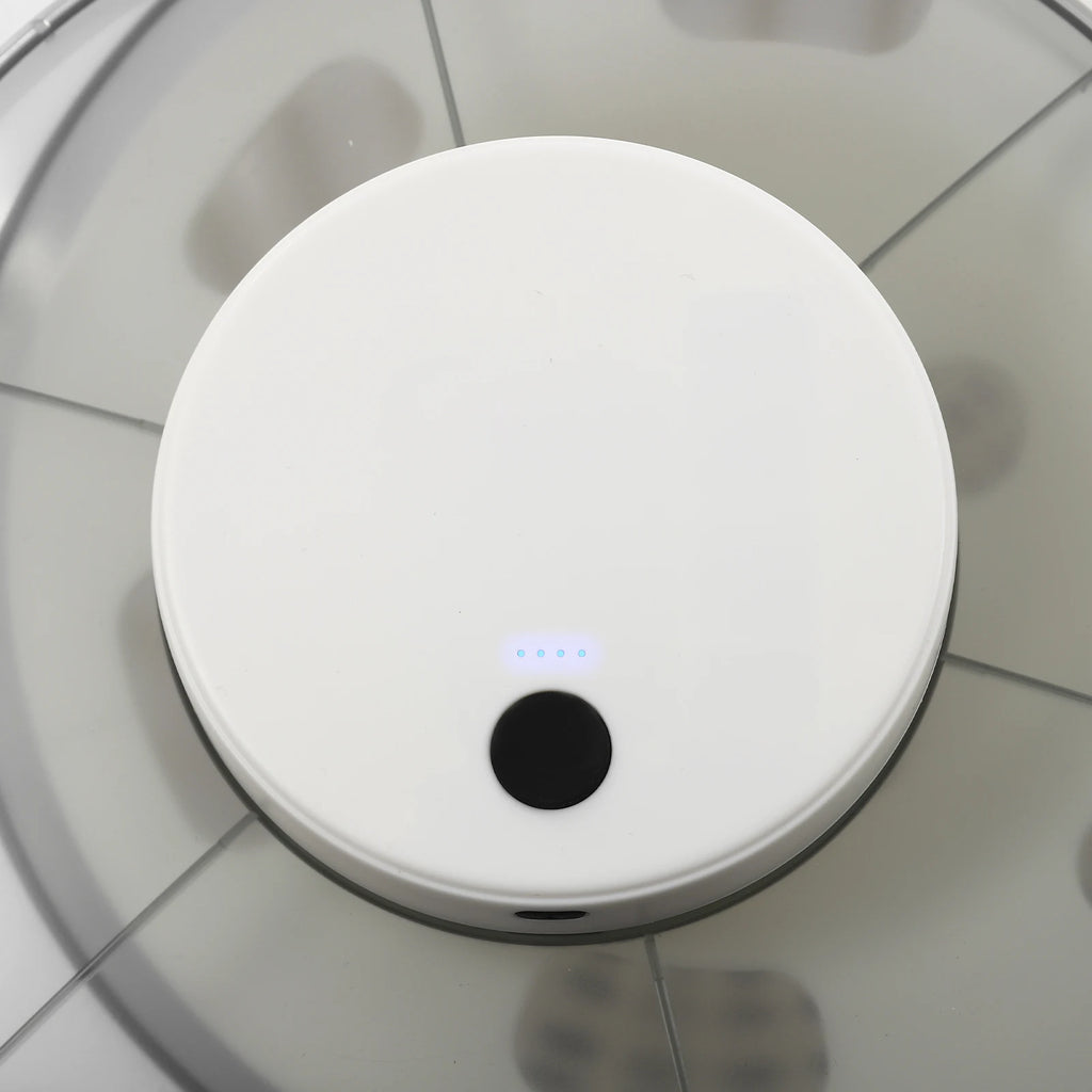 A top-down view of a white round robotic vacuum cleaner on a light gray tiled floor, with a small blue LED indicator light illuminated near the center.