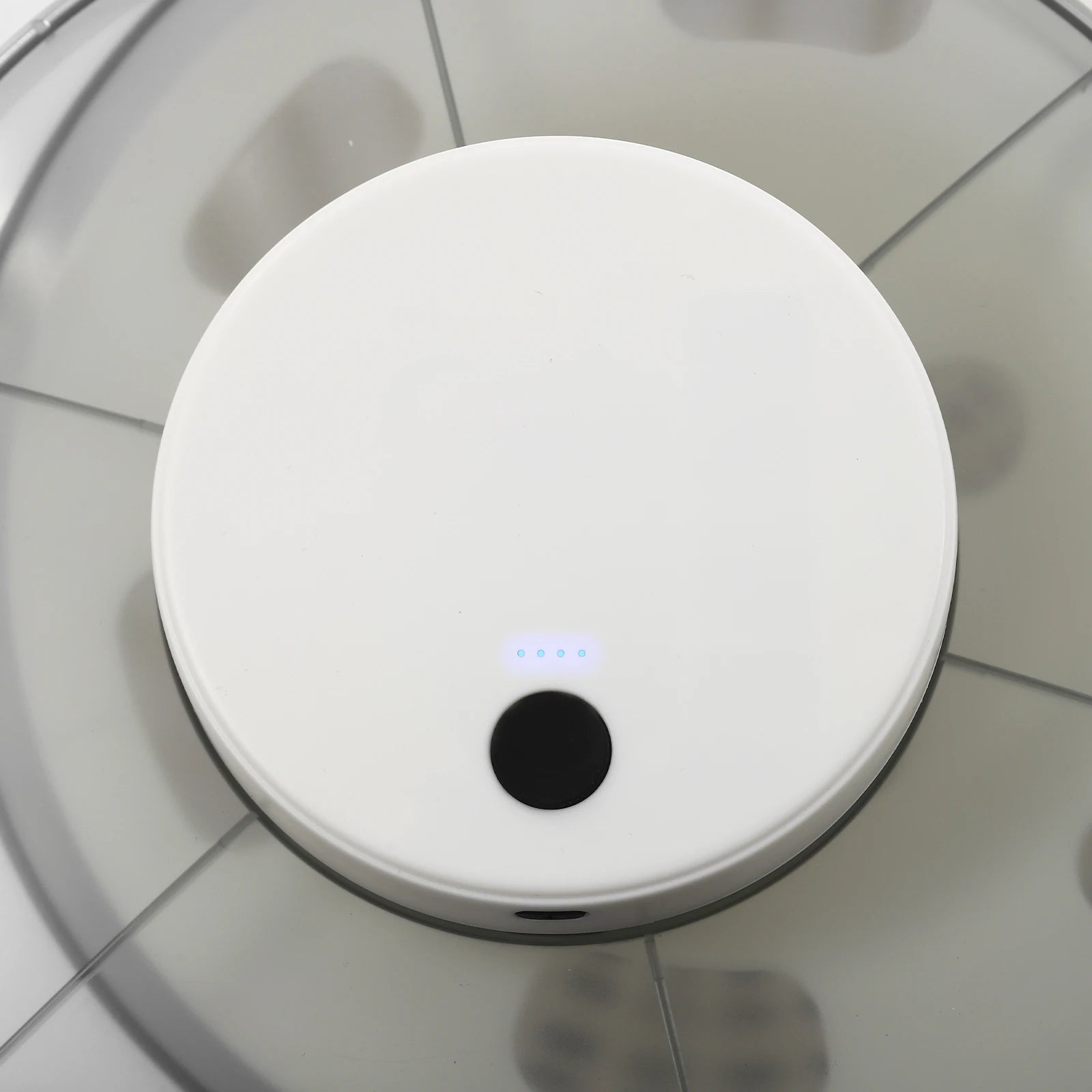 A top-down view of a white round robotic vacuum cleaner on a light gray tiled floor, with a small blue LED indicator light illuminated near the center.