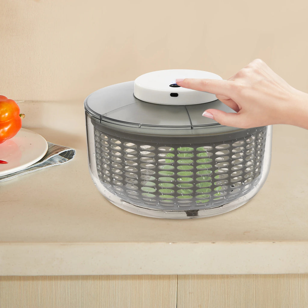A countertop salad spinner with a clear plastic container, gray inner basket, and a white top button being pressed by a person's hand. The scene is set on a beige kitchen counter with a blurred orange fruit on the left.