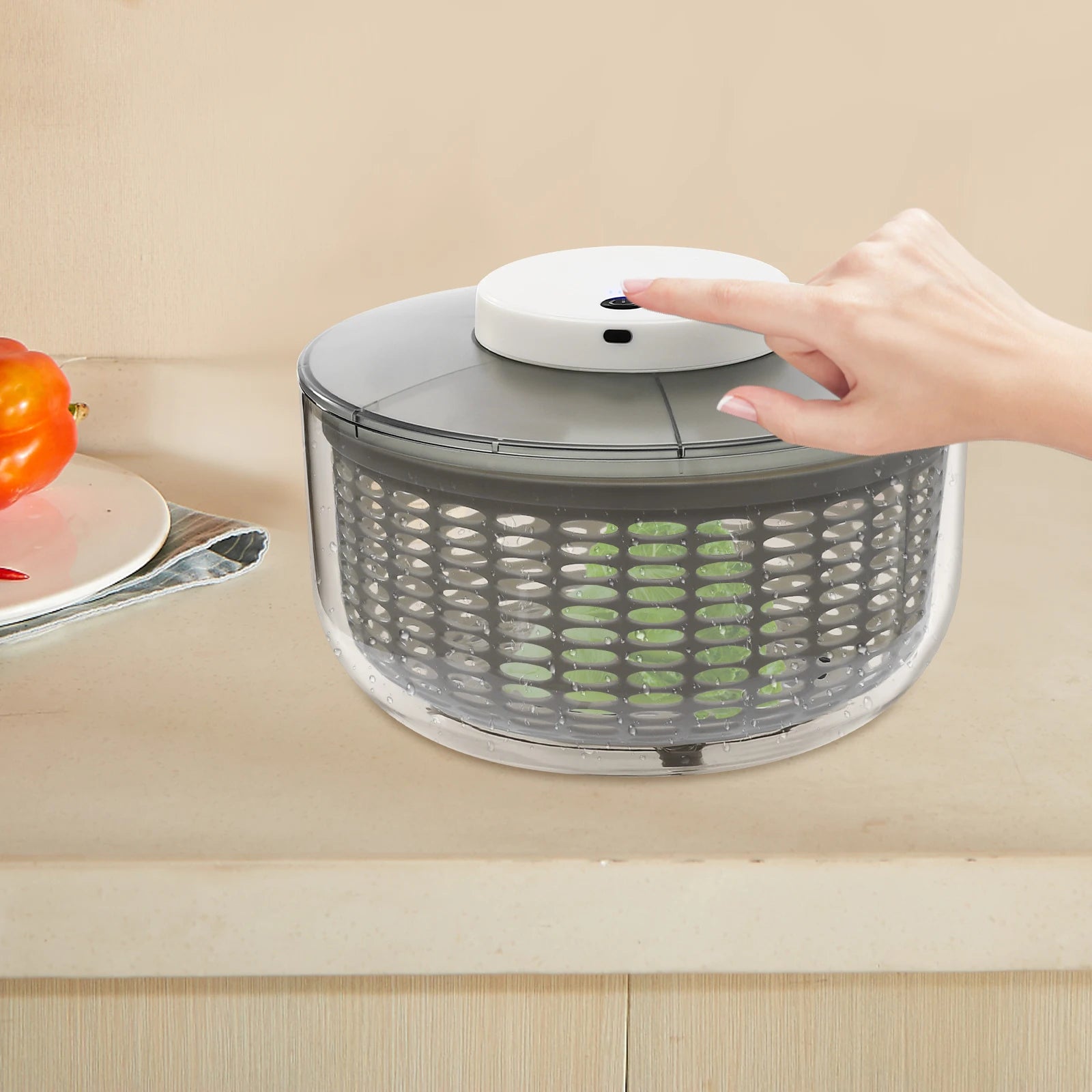 A countertop salad spinner with a clear plastic container, gray inner basket, and a white top button being pressed by a person's hand. The scene is set on a beige kitchen counter with a blurred orange fruit on the left.