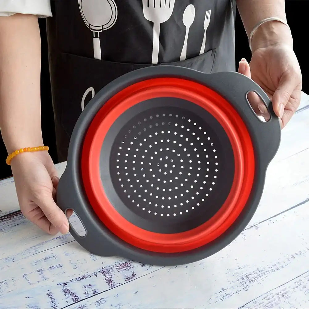 Collapsible silicone strainer with red interior held by a person wearing a black apron with white utensil designs.