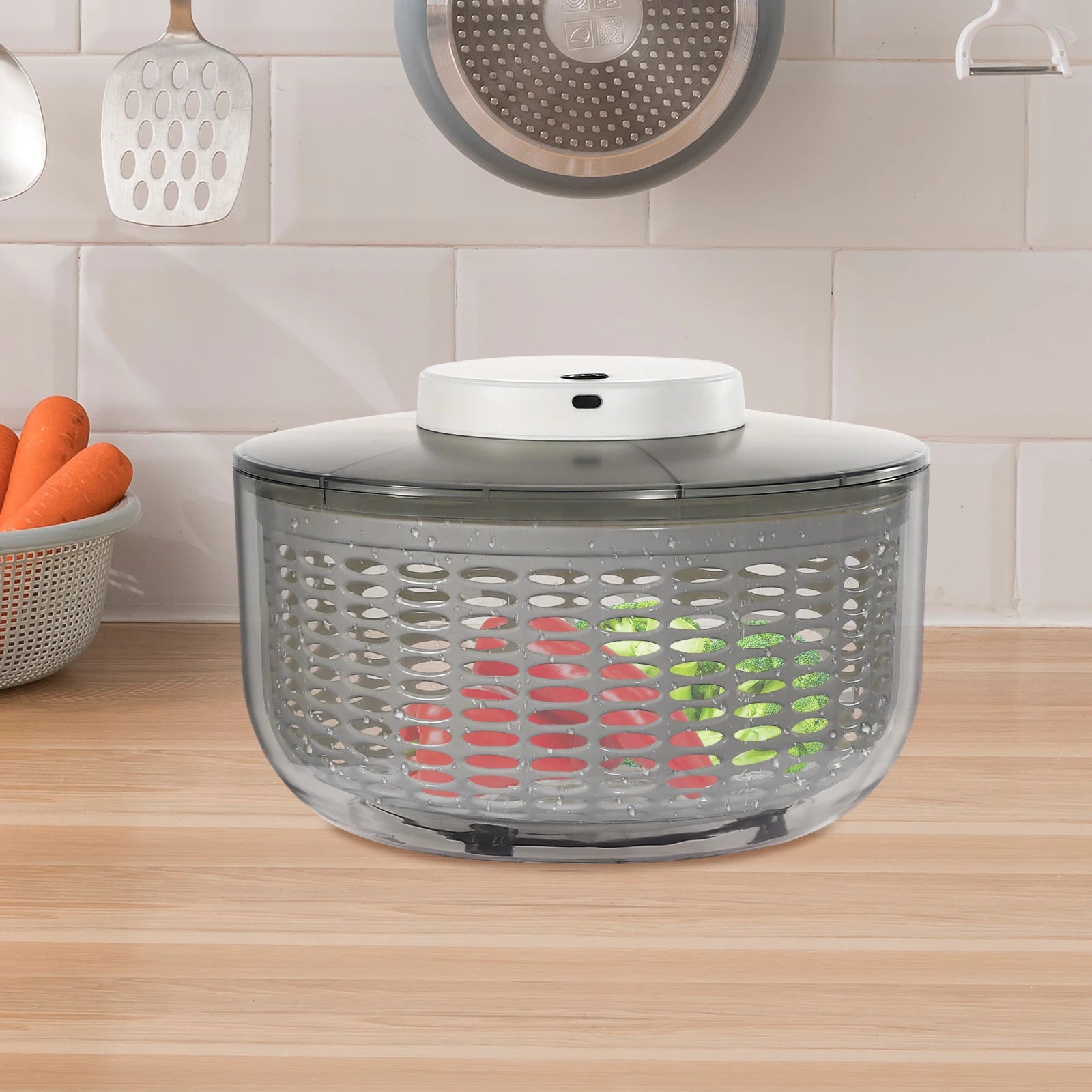 A countertop salad spinner with a clear plastic basket, dark gray lid, and white top button, shown on a wooden kitchen counter with vegetables visible inside.