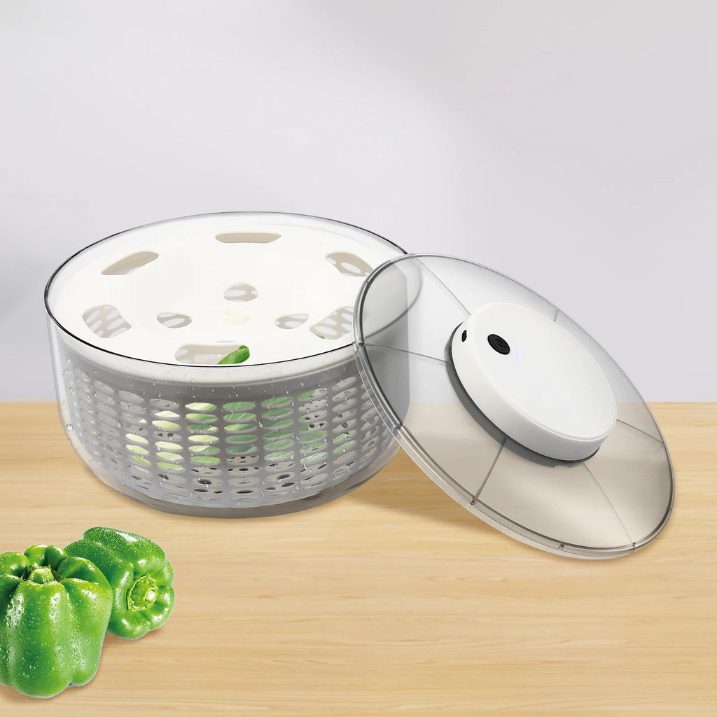 A clear plastic salad spinner basket with a white inner plate and a matching clear lid resting beside it on a light wood surface. Two green bell peppers are placed in the lower left corner.